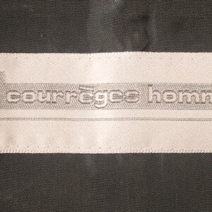 May include: A rectangular white label with the text "courr&egrave;ges homme" in gray, stitched onto a dark gray fabric. The label features a stylized "c" logo.