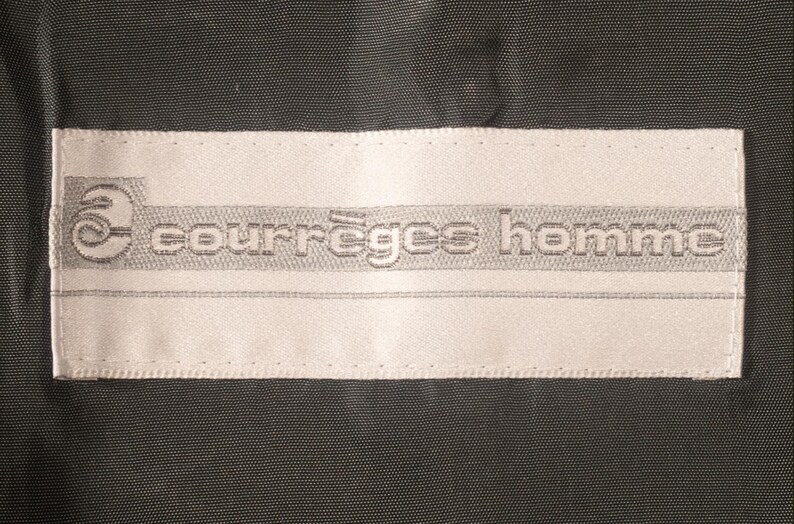 May include: A rectangular white label with the text "courr&egrave;ges homme" in gray, stitched onto a dark gray fabric. The label features a stylized "c" logo.