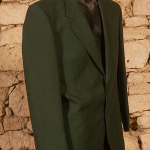 May include: A dark green blazer with a two-button closure and notched lapels. The blazer has three buttons on each cuff and two front pockets. The jacket is displayed on a black mannequin against a stone wall background.