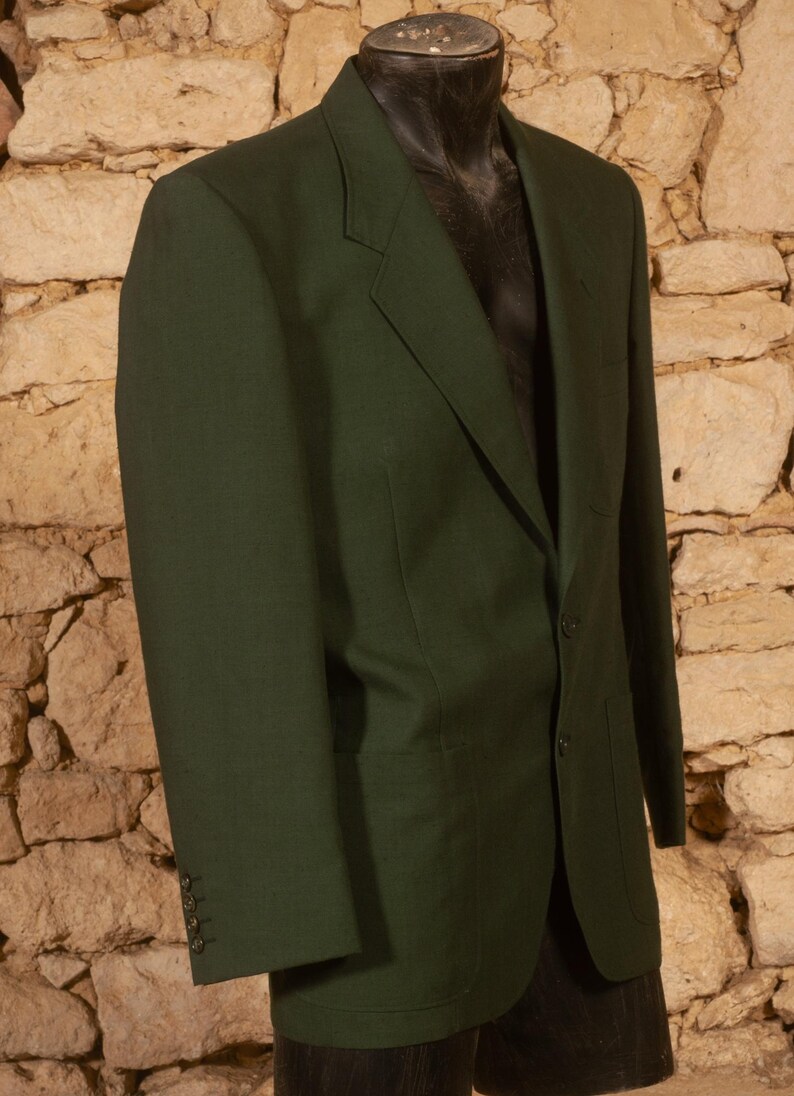 May include: A dark green blazer with a two-button closure and notched lapels. The blazer has three buttons on each cuff and two front pockets. The jacket is displayed on a black mannequin against a stone wall background.