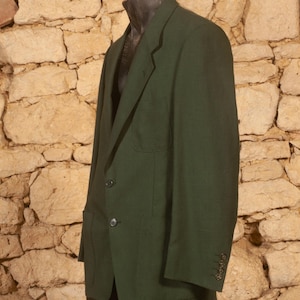 May include: A dark green blazer with two front pockets and three buttons on each cuff. The blazer is displayed on a mannequin against a textured stone wall background. The jacket has a two-button closure.