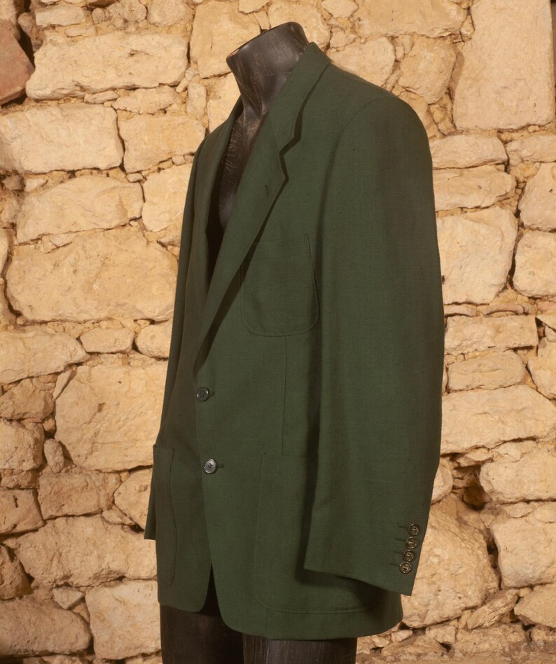 May include: A dark green blazer with two front pockets and three buttons on each cuff. The blazer is displayed on a mannequin against a textured stone wall background. The jacket has a two-button closure.