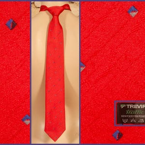 Peut inclure: Une cravate rouge vif présentant un motif texturé diagonal subtil et de petits accents en forme de losange bleu et gris régulièrement espacés. La cravate comprend une étiquette noire avec le texte Trevira traffic 100% polyester/poliestere.