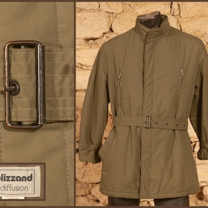 Puede incluir: Una chaqueta de color verde oliva con cuello alto, mangas largas y un cinturón a juego con una hebilla metálica rectangular. La chaqueta incluye dos bolsillos verticales con cremallera en el pecho y una etiqueta blizzand diffusion.