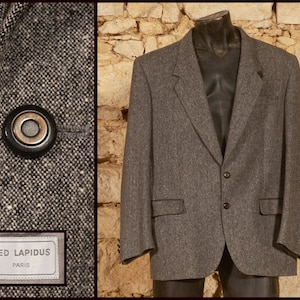 Op de afbeelding: Een grijze tweed blazer met een sluiting met twee knopen, inkepingen in de revers en twee klepzakken. Het kledingstuk bevat een label met de tekst TED LAPIDUS PARIS en een close-up van een zwarte en gouden knoop op een textuurstof.