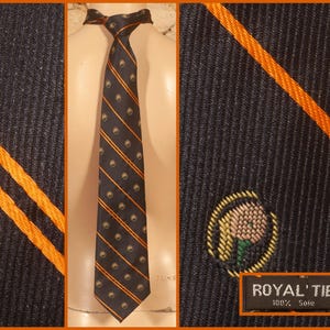 Peut inclure: Une cravate noire à texture côtelée ornée de rayures diagonales orange et de petits emblèmes circulaires brodés. La cravate comporte une étiquette noire avec le texte blanc ROYAL' TIE 100% Soie, indiquant la composition du matériau.