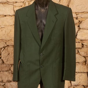 May include: A dark green blazer with two front pockets, a notched lapel, and two-button closure. The blazer is displayed on a black mannequin against a textured stone wall background. The blazer has a classic, tailored design.