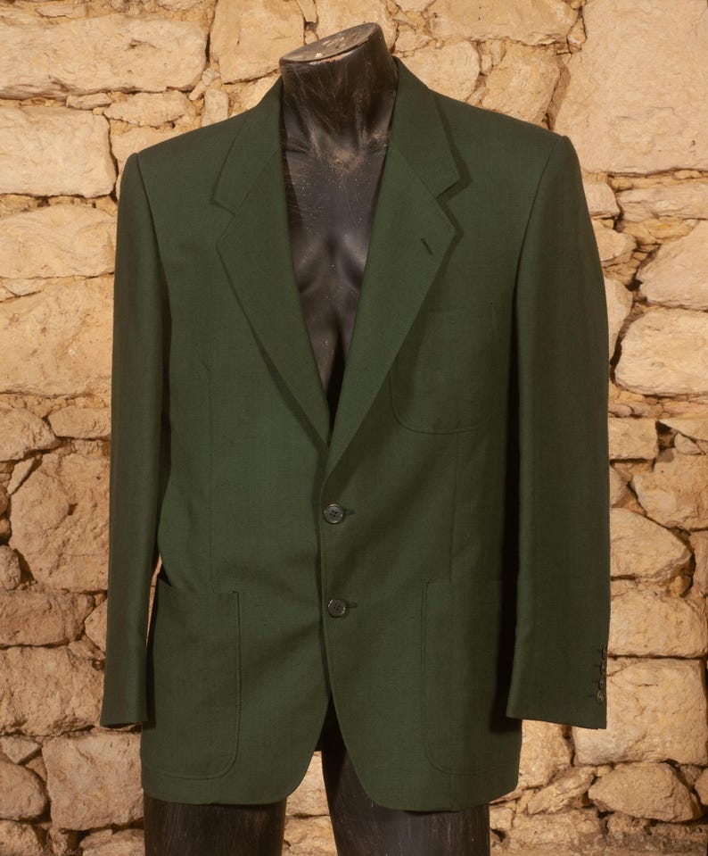 May include: A dark green blazer with two front pockets, a notched lapel, and two-button closure. The blazer is displayed on a black mannequin against a textured stone wall background. The blazer has a classic, tailored design.
