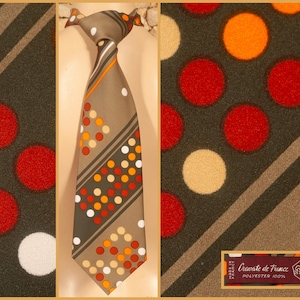 May include: A brown necktie featuring a geometric pattern of diagonal stripes and circular dots in shades of red, orange, and cream. The tie includes a label that reads "Cravate de France" and "POLYESTER 100%" with "MADE IN FRANCE" text.