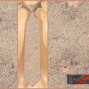 May include: A necktie in a muted taupe colour featuring a subtle, repeating floral pattern with small pink blossoms and grey leaves. The fabric has a textured, woven appearance. A dark blue label reads "Pierre J'Espois Paris" and "Made in France".