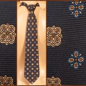 Può includere: Cravatta blu navy con un motivo ripetuto di disegni floreali e quadrati nei toni del marrone, beige e blu. La cravatta è esposta su un manichino, mostrando la sua lunghezza e il nodo.