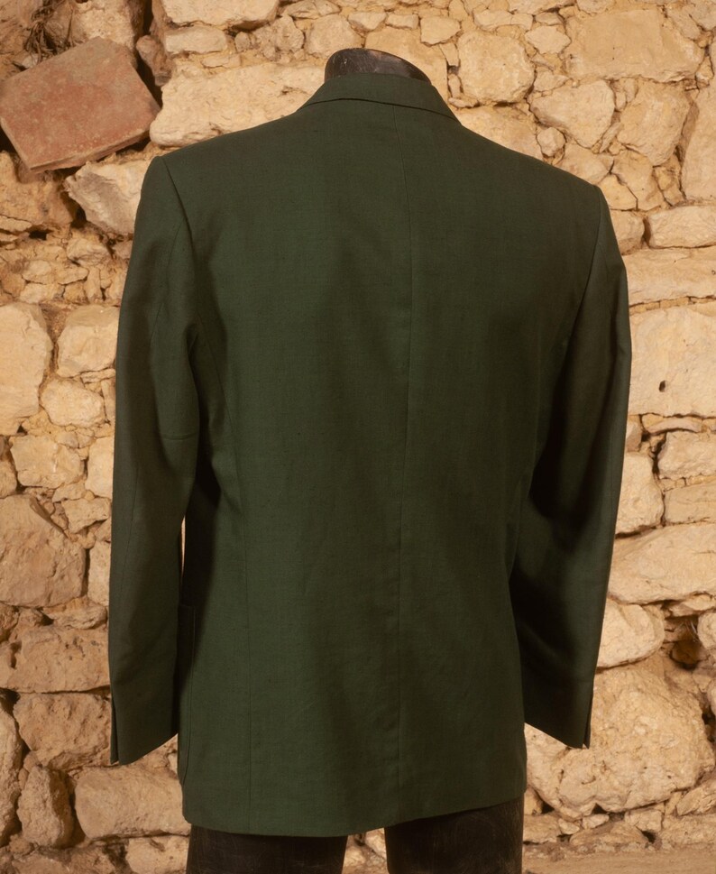 May include: A dark green blazer with a two-button closure, shown from the back. The blazer has a tailored fit and a classic design. The fabric appears to be a solid color, with a subtle texture. The blazer is displayed on a mannequin.