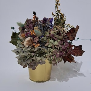May include: A dried flower arrangement in a gold metal bucket. The bouquet features a variety of dried flowers in shades of purple, pink, green, and yellow.