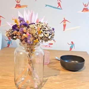 Könnte beinhalten: Eine klare Glasvase mit einem kupferfarbenen geometrischen Muster ist mit getrockneten Blumen gefüllt. Die Vase steht auf einem Holztisch mit einer schwarzen Schüssel und rosa Untersetzern.