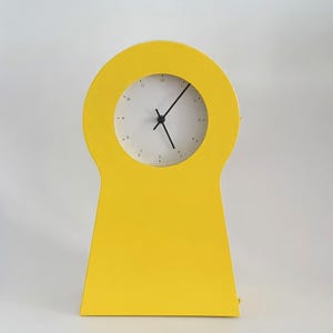 May include: A yellow keyhole-shaped clock with a white face and black hands. The clock is on a white background.