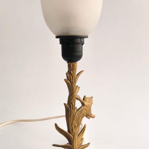 Op de afbeelding: Een vintage tafellamp met een witte glazen kap met een bloemmotief. De lamp heeft een messingkleurige voet met een gedetailleerd botanisch motief en een zwarte lamphouder. De lamp is ongeveer 30 cm hoog.