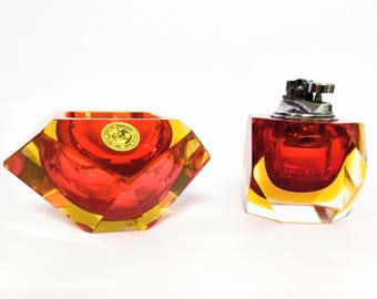 May include: A pair of vintage Murano glass objects. One is a faceted ashtray in red and yellow. The other is a matching lighter with a silver-tone metal top. Both have a geometric design.