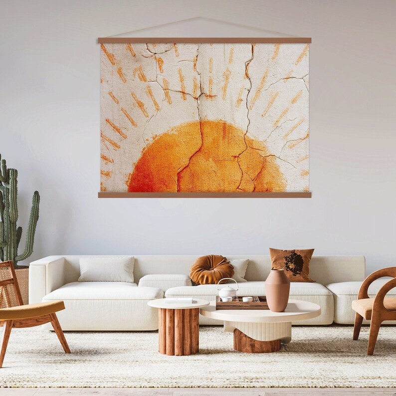 May include: A large wall hanging featuring an abstract design of a sun. The sun is a vibrant orange color, set against a textured, off-white background with radiating lines. The artwork is framed by a wooden hanging system.