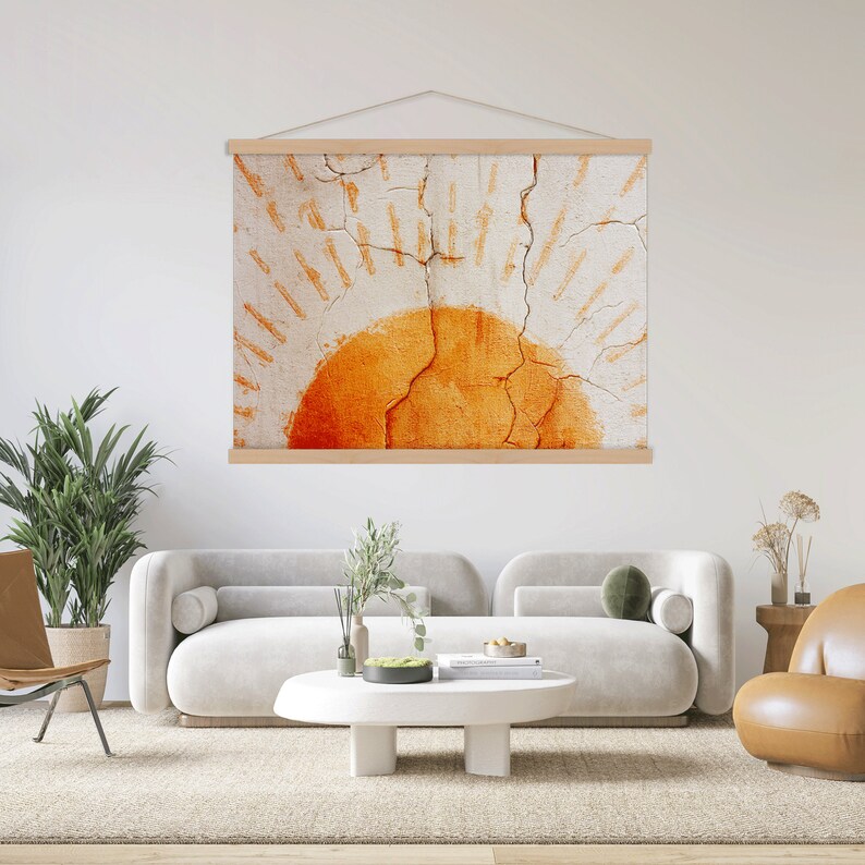 May include: A wall hanging with a sun design. The artwork features a textured, off-white background with an orange sunburst design. The artwork is held by a natural wood frame and suspended by a white cord. The piece is displayed in a modern living room.