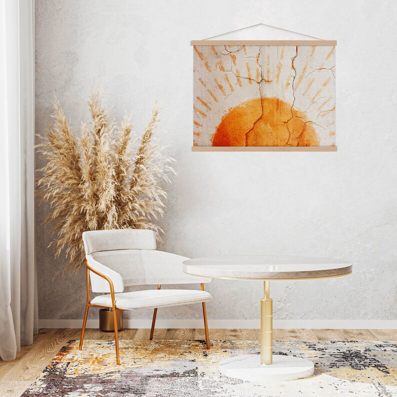 May include: A framed wall art print featuring an abstract design of a sun in shades of orange and beige. The artwork is displayed in a light wood frame, hanging above a white table with a gold base. A beige chair and decorative dried plants complete the scene.