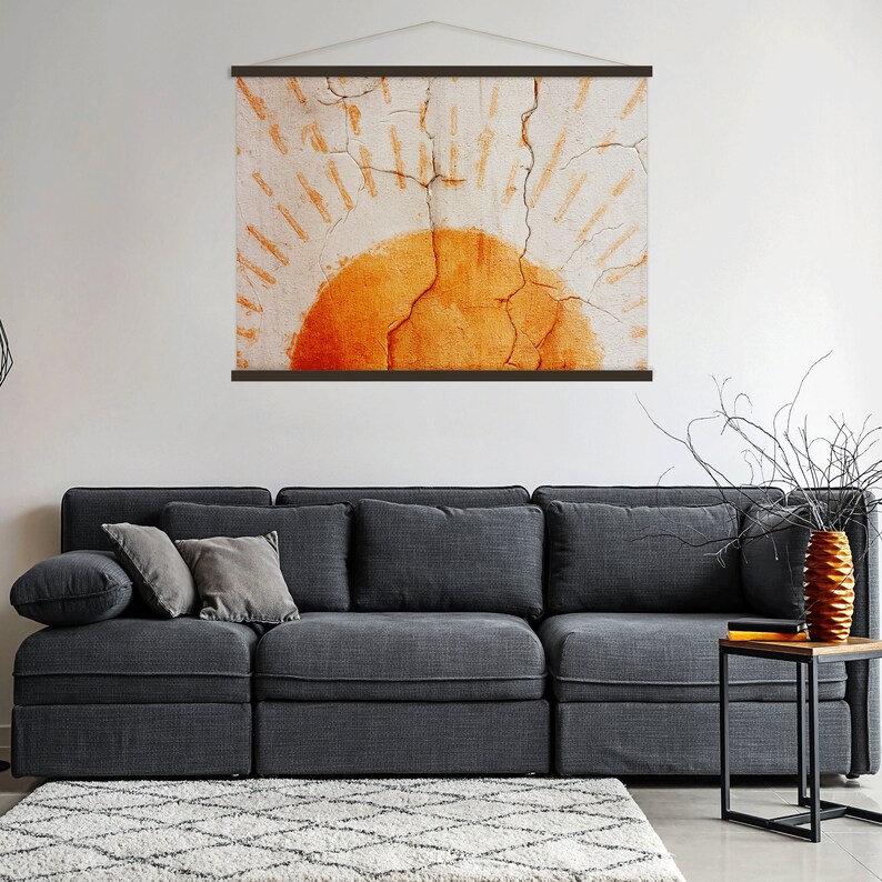 May include: A wall hanging featuring an abstract sun design in shades of orange and cream. The artwork is mounted with a dark brown wooden frame and a natural cord for hanging. The sun design has a textured, cracked appearance.