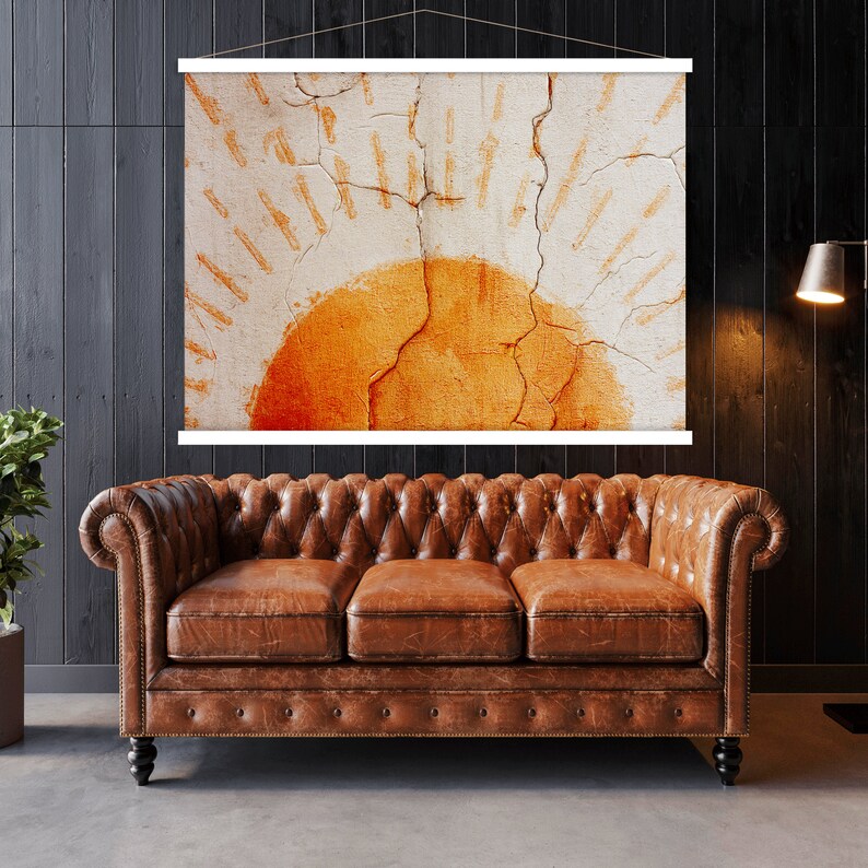 May include: A framed wall art print featuring an abstract sun design in shades of orange and white. The artwork is displayed above a brown leather Chesterfield sofa. The room has dark wood paneling and a floor lamp.
