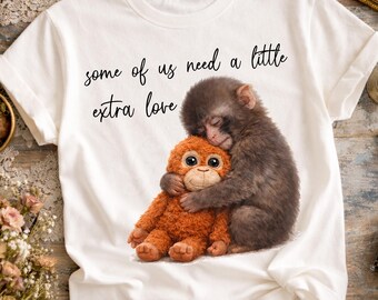 May include: White t-shirt with a graphic of a monkey cuddling an orange soft toy. The text "some of us need a little extra love" is written above the graphic in black script. Digital download.