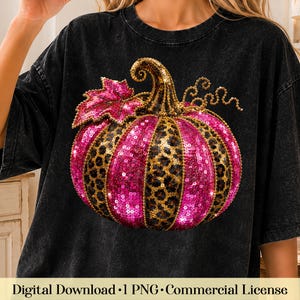 May include: A black acid-wash t-shirt featuring a large graphic of a pumpkin. The pumpkin is decorated with alternating vertical panels of bright pink sequins and a brown and black leopard print pattern, topped with a single pink sequin leaf.