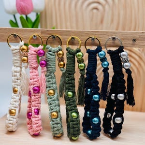 Könnte beinhalten: Eine Sammlung handgefertigter Makramee-Schlüsselanhänger in verschiedenen Farben, darunter Creme, Rosa, Grün, Marineblau und Schwarz. Jeder Schlüsselanhänger hat dekorative Perlen in Gold, Rosa, Grün, Blau und Silber und ist an einem goldfarbenen Metallring befestigt.