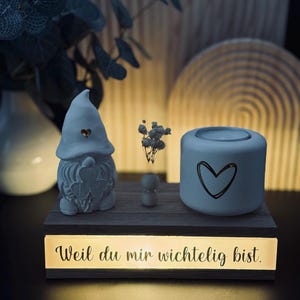 Könnte beinhalten: Dekorative Holz-Anzeige mit beleuchtetem Sockel. Sie verfügt über eine Zwergfigur, einen kleinen Kerzenhalter mit Herzdesign und den Text "Weil du mir wichtig bist." Die Anzeige wird von einem warmen Schein beleuchtet.