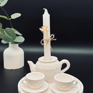 Könnte beinhalten: Weißer Keramikkännchen-Kerzenhalter mit zwei kleinen Teetassen und Untertassen auf einem weißen Tablett. Die Teekanne hat ein dekoratives Design und die Tassen haben goldene Herzen auf der Vorderseite.