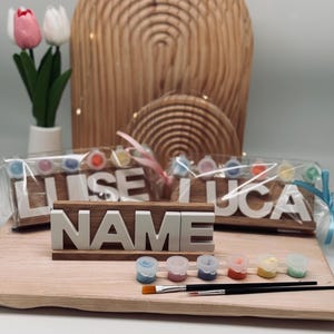 Könnte beinhalten: Ein Holzschild mit dem Wort "NAME" in weißen Blockbuchstaben. Das Schild steht auf einem Holzsockel, mit Farbtöpfen und Pinseln. Zwei weitere Holzschilder mit Namen sind im Hintergrund, zusammen mit Farbtöpfen.