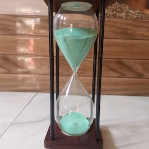 Könnte beinhalten: Eine Sanduhr aus Holz mit einem schwarzen Metallrahmen und grünem Sand. Die Sanduhr steht auf einer weißen Oberfläche.