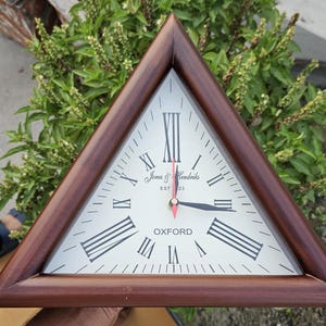 May include: A triangular wooden clock with a white face and black hands. The clock face has Roman numerals and the text "Jones & Hendricks EST 1923 OXFORD".