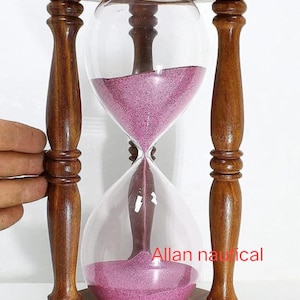 Può includere: Una clessidra di legno con una clessidra di sabbia rosa. La clessidra è realizzata in legno scuro con una finitura lucida. La clessidra è alta circa 25 cm.