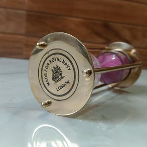 May include: Brass hourglass with a pink sand timer and a brass base that reads "Made for Royal Navy London".