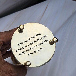 May include: A brass hourglass with a circular base and top. The top is engraved with the words "This sand and this hourglass symbolize our unity and love until the end of time."