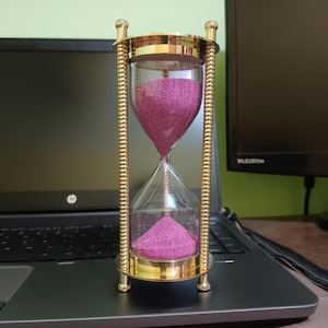 May include: An hourglass with a gold-colored metal frame and clear glass chambers. The sand inside is a vibrant magenta color. The hourglass is sitting on a laptop.