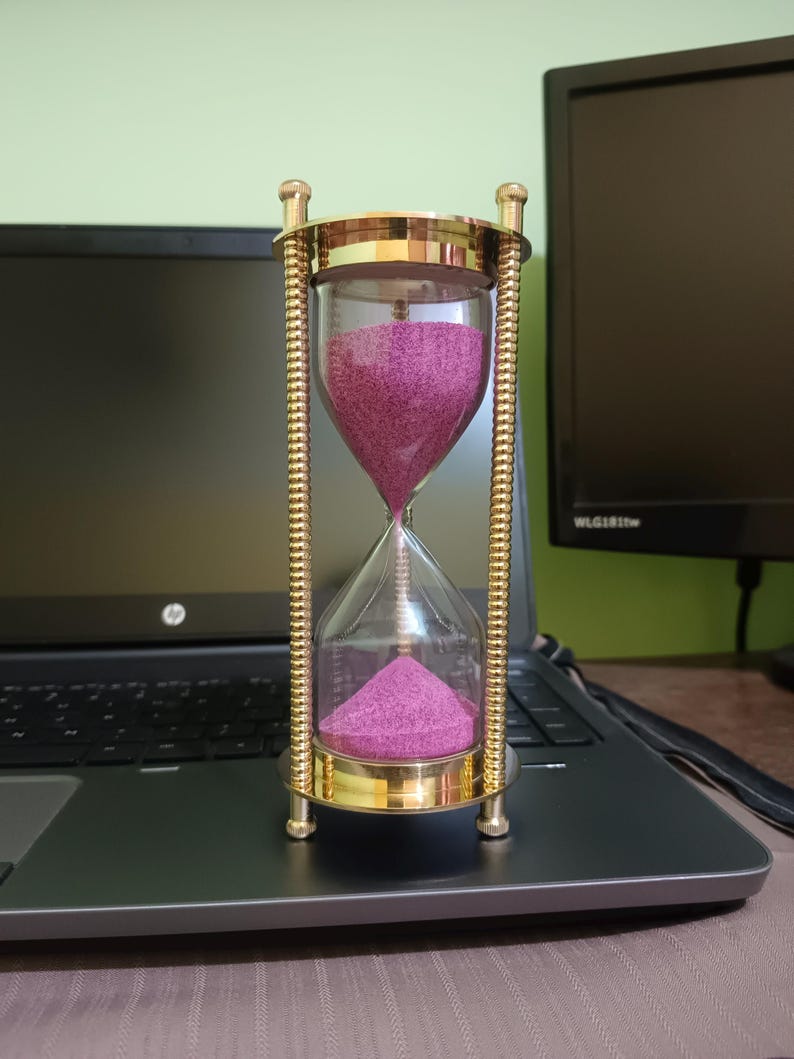 May include: An hourglass with a gold-colored metal frame and clear glass chambers. The sand inside is a vibrant magenta color. The hourglass is sitting on a laptop.