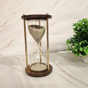 以下が含まれることがあります： 木製のフレームとベースを備えた砂時計で、透明なガラスと金色の金属製のサポートが特徴です。砂時計には明るい色の砂が入っています。木製の要素は濃い茶色の仕上げです。装飾品です。