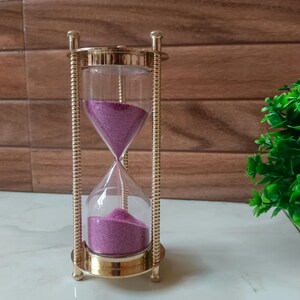 May include: An hourglass with a gold-colored metal frame and clear glass containers. The sand inside is a vibrant purple color. The hourglass is a decorative item, likely used for timing or as a display piece.