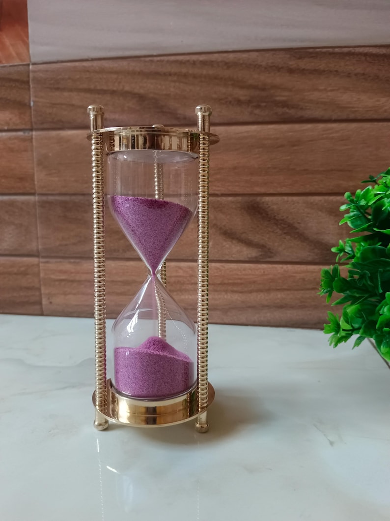 May include: An hourglass with a gold-colored metal frame and clear glass containers. The sand inside is a vibrant purple color. The hourglass is a decorative item, likely used for timing or as a display piece.