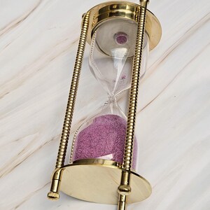 May include: An hourglass with a brass frame and clear glass casing. The sand inside is a vibrant purple color. The hourglass has a classic design with a round base and top.
