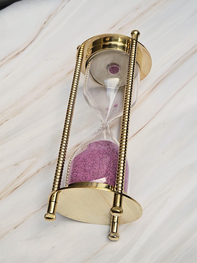 May include: An hourglass with a brass frame and clear glass casing. The sand inside is a vibrant purple color. The hourglass has a classic design with a round base and top.