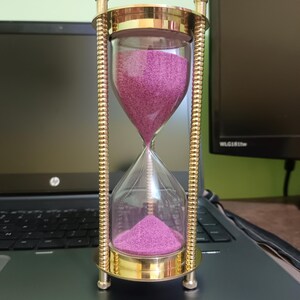 May include: An hourglass with a clear glass body and a brass-colored metal frame. The sand inside is a vibrant magenta color. The hourglass is resting on a laptop, with a monitor in the background.