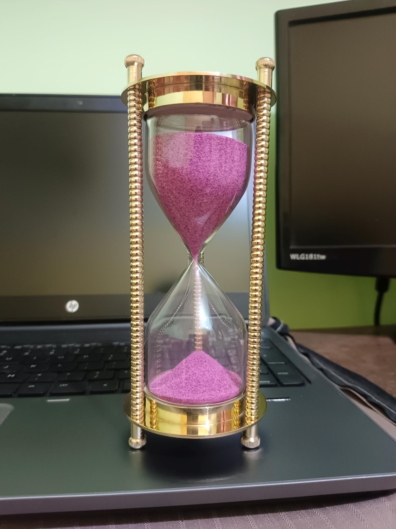 May include: An hourglass with a clear glass body and a brass-colored metal frame. The sand inside is a vibrant magenta color. The hourglass is resting on a laptop, with a monitor in the background.
