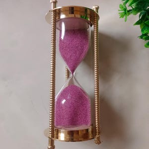 May include: An hourglass with a gold-colored metal frame and clear glass bulbs. The glass bulbs are filled with pink sand. The hourglass is a decorative item, suitable for home decor or as a unique gift.