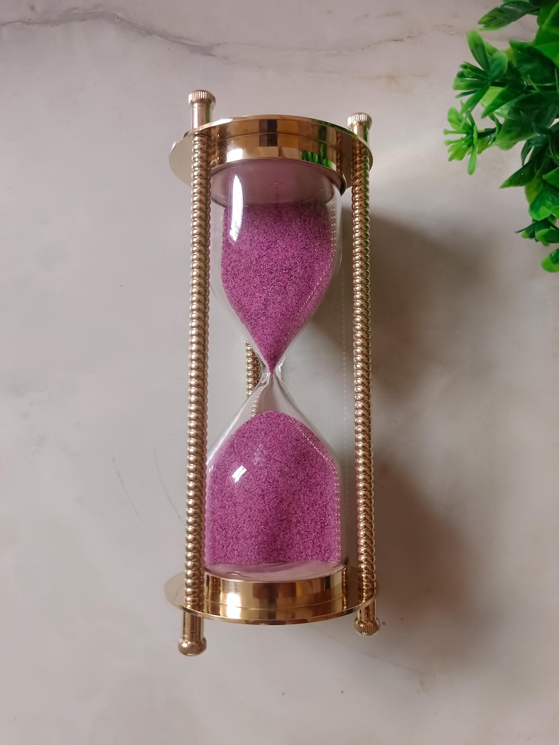 May include: An hourglass with a gold-colored metal frame and clear glass bulbs. The glass bulbs are filled with pink sand. The hourglass is a decorative item, suitable for home decor or as a unique gift.
