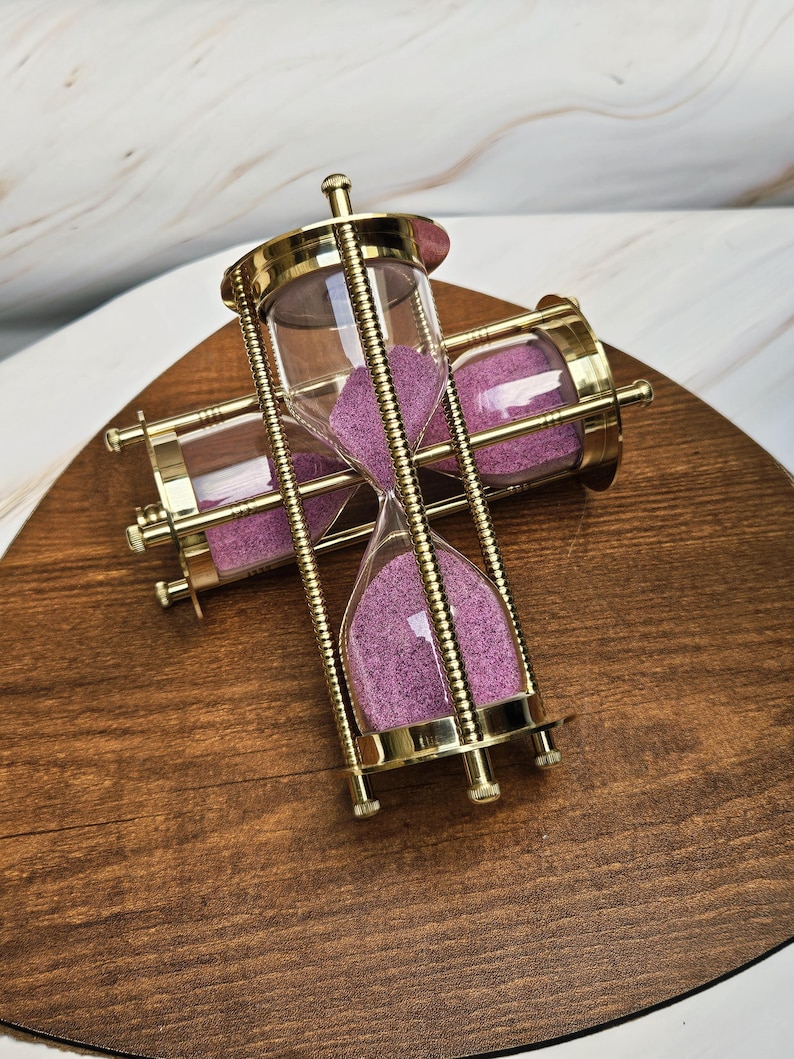May include: Two hourglasses with gold-colored metal frames and clear glass containers filled with purple sand. The hourglasses are positioned on a wooden surface, showcasing their decorative design and the contrast of colors.