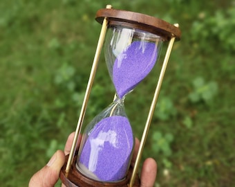 May include: Hourglass with a wooden frame and brass supports, holding purple sand. The hourglass has a clear glass bulb and a dark brown wooden base. The background is a blurred green.